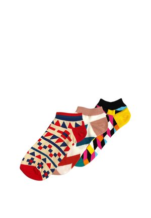 Jeune femme avec des tresses blondes et des créoles dorées assise en tailleur, vêtue d’un t-shirt jaune et de chaussettes à motifs rouges, bleus et beiges, souriante.