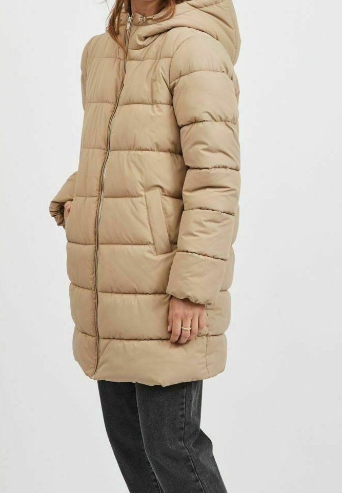 Veste matelassée longue beige avec capuche, fermeture éclair frontale et poches latérales portée avec un pantalon noir.
