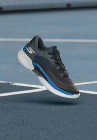 Schwarzer Sportschuh mit blauen Akzenten, Mesh-Obermaterial, runder Zehenpartie, strukturierten Details und weißer Außensohle, erhöht über einer blauen Platzoberfläche.