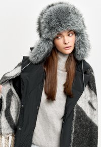 Căciulă gri din blană sintetică cu urechi, asortată cu o jachetă neagră din puf și un pulover gri cu dungi, purtând un șal cu model țesut.