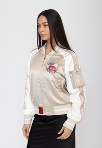 Frau trägt beige-weiße Satin-Bomberjacke mit floraler Stickerei auf der Brust und Reißverschlusstasche am Ärmel, kombiniert mit schwarzem Rock.