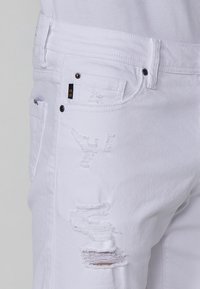 Jeans de mezclilla blanca con acentuaciones desgastadas, dobladillos deshilachados y costuras visibles. Incluyen dos bolsillos y una etiqueta negra cerca de la cinturilla.