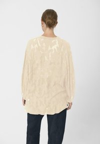 Šviesiai smėlinė didelio dydžio bliuska su tekstūruotu gėlių raštu, laisvais rankovėmis ir apvaliu žemyn kraštu, derinama su tamsiai džinsinėmis kelnių.