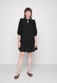 Veste preta com mangas balão 3/4, decote em gota e bolinhas brancas. Base de camadas esvoaçante, feita de tecido leve. Sandálias pretas.