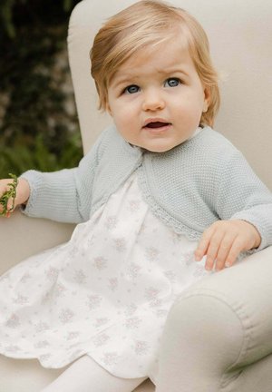 Hellblau gestrickte Strickjacke über einem weißen Kleid mit kleinen Blumenmustern. Kind sitzt auf einem beigen Stuhl und hält grüne Blätter.