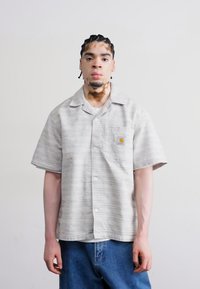 Kurzärmliges, knöpfbares Hemd in Hellgrau mit einem subtilen Streifenmuster. Mit einer Brusttasche und einem Logodetail aus Stoff. Kombiniert mit blauen Jeans.