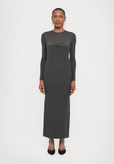 Robe ajustée à manches longues en tissu gris foncé. Elle présente un panneau supérieur transparent et des fentes sur les côtés. Une paire d'escarpins noirs à bout pointu est portée.