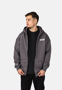 Felpa grigia con zip, dotata di tasca frontale, polsini a coste e un logo bianco con la scritta "CRYSTAL". Indossata sopra una t-shirt nera.