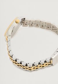 Rozsdamentes acél karóra arany akcentussal; szegmensekből álló karkötő dizájnja van, váltakozó fényes ezüst és arany láncszemekkel, a korona 2 órakor található.