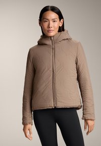 Brun quiltad jacka med huva, med dragkedja framtill och strukturerat mönster. Hemmets och ärmarna har en mjuk, fluffig kant. Bärs över svarta leggings.