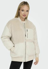 Giacca beige chiaro con una parte superiore morbida, polsini elasticizzati e una tasca con zip frontale. Il design combina materiali lisci e testurizzati.