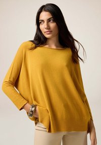 Pull oversized jaune moutarde avec des manches longues côtelées et un ourlet. Présente des poches sur les côtés. Porté avec un bracelet à motifs. Texture douce.