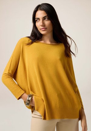 Senfgelbes, übergroßes Sweater mit gerippten, langen Ärmeln und Saum. Verfügt über seitliche Taschen. Tragen mit einem gemusterten Armreif. Weicher Stoff.