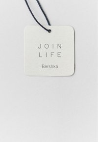 Etiqueta de tela blanca con esquinas redondeadas, que presenta el texto "JOIN LIFE" en gris y "Bershka" abajo, sujeta con un cordón negro.