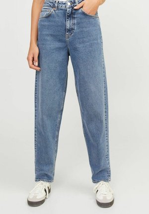 Personne portant un jean bleu ample et des baskets blanches avec des détails noirs, debout avec une main dans la poche devant un fond uni.