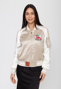 Frau mit langen dunklen Haaren, die eine beige-weiße Satin-Bomberjacke mit Blumenstickerei und einen schwarzen Rock vor einem einfarbigen Hintergrund trägt.