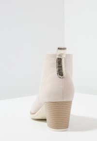 Stivaletto in suede beige con un tassello metallico dorato, tacco quadrato testurizzato e dettagli di cucitura sul retro e sui lati.