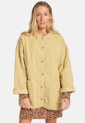 Gepolsterte gelbe Jacke mit rundem Ausschnitt, vorderen Knöpfen und zwei Taschen; getragen über einem blumigen braunen Kleid mit langen Ärmeln.