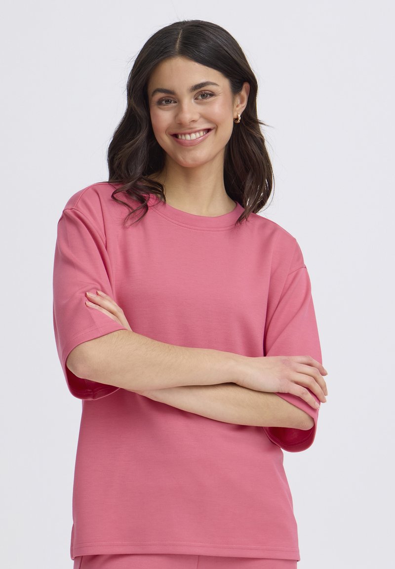 Ung kvinde med mørkt, bølget hår iført en løs pink T-shirt, smilende med armene krydset foran en ensfarvet lys grå baggrund.