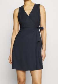 Bezramenné námořnické modré zavinovací šaty s V-výstřihem, uvázané v pase, nošené osobou stojící na jednobarevném pozadí.