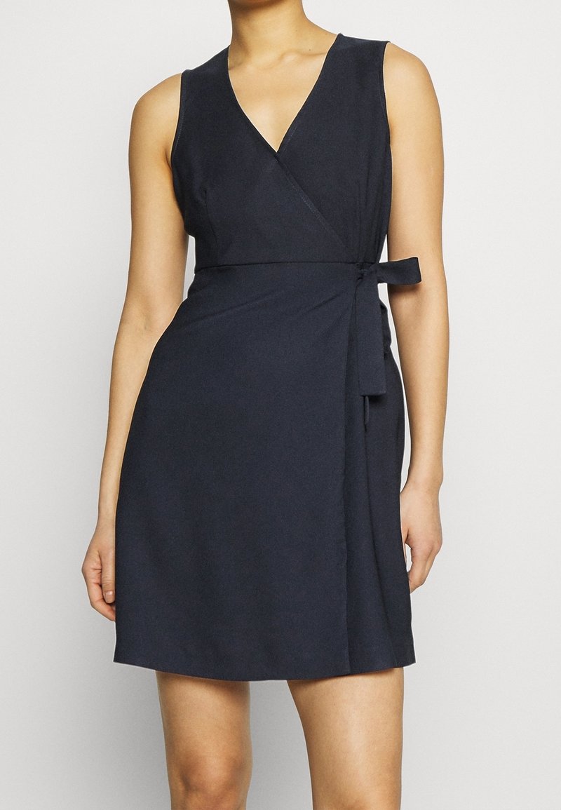 Bezramenné námořnické modré zavinovací šaty s V-výstřihem, uvázané v pase, nošené osobou stojící na jednobarevném pozadí.