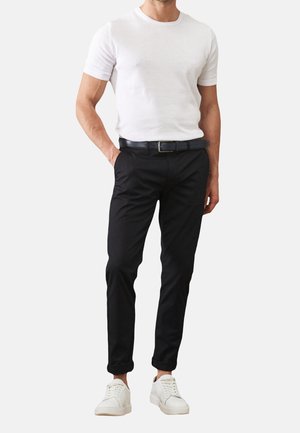 Man draagt een wit T-shirt met korte mouwen, zwarte broek met een zwarte riem, en witte sneakers, staand met één hand in de zak.