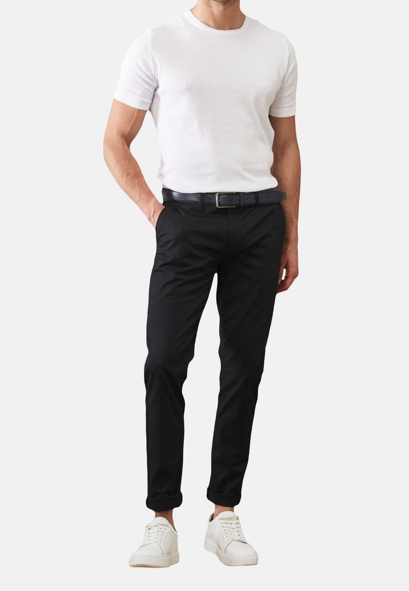 Homme portant un t-shirt blanc à manches courtes, un pantalon noir avec une ceinture noire et des baskets blanches, debout avec une main dans la poche.