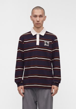Mand iført en mørkeblå polo med lange ærmer med hvide, orange og beige vandrette striber og grå bukser, der står mod en ensfarvet baggrund.