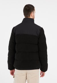 Sort pufferjakke med en struktureret fleece-krop og glatte stofskuldre, med en høj krave og elastisk kant, i en afslappet pasform.