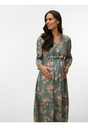 Femme enceinte souriante regardant sur le côté, portant une robe de maternité verte à fleurs à manches longues, les mains posées doucement sur son ventre.