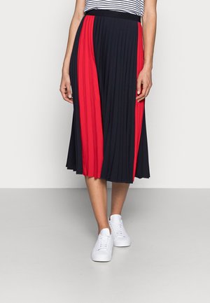 Femme portant une jupe midi plissée avec des panneaux verticaux rouges et noirs, des baskets blanches, et une partie d'un haut rayé visible.