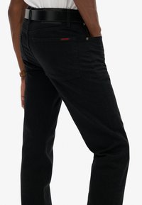 Jean en denim noir avec une coupe slim, des poches arrière et une étiquette rouge "SUPERDRY". Le tissu présente des coutures visibles et une texture douce.