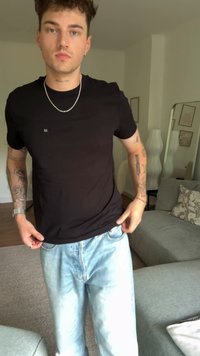 Jaunas vyras su trumpais garbanotais plaukais, apsirengęs juodu marškinėliu ir šviesiai mėlynomis džinsomis, stovi svetainėje, turintis tatuiruotes ant abiejų rankų.
