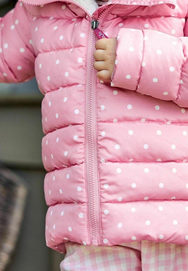 Enfant portant une doudoune rose à pois avec une main visible qui tient la fermeture éclair avant.