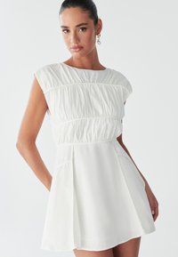 Hvid kort kjole med en tætsiddende krop med rynkede syninger, kasketærmer og en udvidet nederdel. Inkluderer sidelommer for funktionalitet.