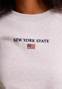 Detailní záběr na světle šedou košili s vyšitým nápisem "NEW YORK STATE" nad malou americkou vlajkou.