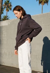 Jeune femme portant un sweat à capuche sombre et un pantalon blanc, debout à l'extérieur contre un mur gris avec des palmiers en arrière-plan.