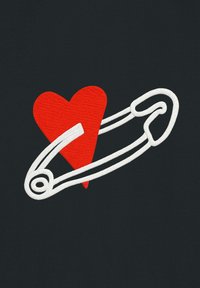 Un tissu noir arbore un grand cœur rouge et un motif d'épingle de sûreté blanche, tous deux réalisés avec une broderie texturée.