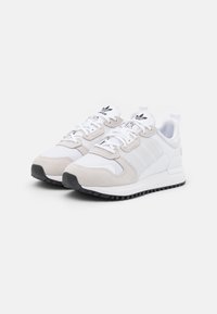 Bílé atletické tenisky se šedými semišovými doplňky a síťovanými panely. Mají tři bílé pruhy a černou gumovou podrážku s texturovaným gripem.