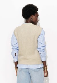 Gilet en tricot beige avec un col rond, porté sur une chemise à rayures bleues avec des détails de boutons, associé à un jean bleu clair et des bracelets en or.