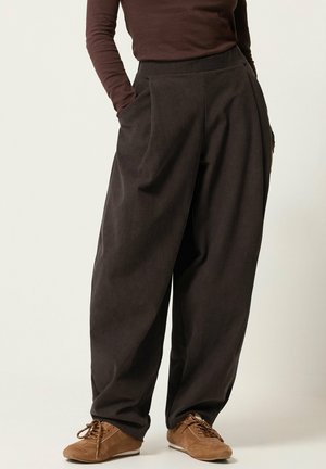Persona che indossa pantaloni larghi di velluto a coste marrone scuro e scarpe stringate in camoscio marrone, in piedi con le mani nelle tasche su sfondo chiaro.