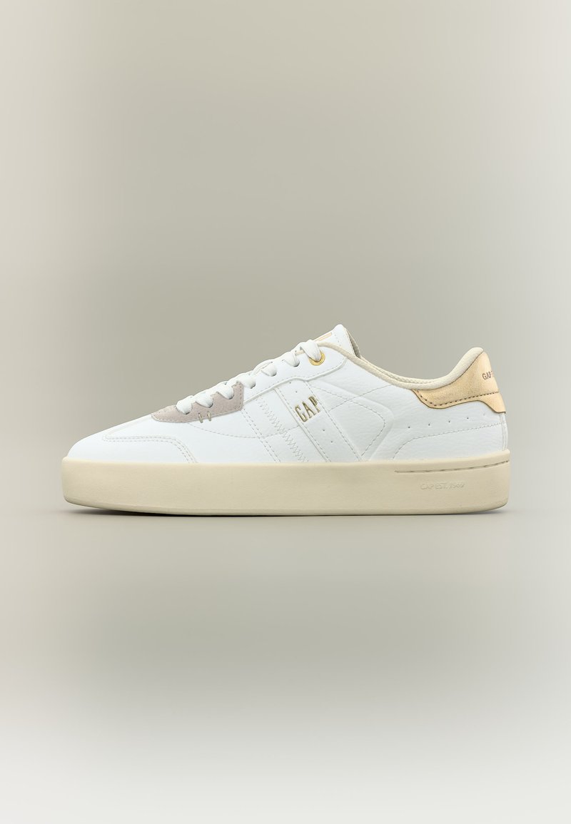 Baskets basses blanches avec un onglet de talon en daim beige, une semelle crème, des lacets blancs et le logo "GAP" sur le côté et le talon.