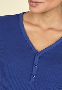 Sinine ribaga topp, millel on V-kujuline kaelus ja kolm lillat nööpi ees. Kangas näeb välja pehme ja veidi tekstuurne.