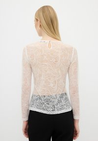 Maglietta a maniche lunghe in pizzo di un beige chiaro con motivi floreali e una texture trasparente, caratterizzata da un orlo inferiore in pizzo nero e un piccolo foro a forma di chiave sul retro.
