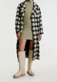 Manteau à carreaux en noir et blanc, robe en vert olive côtelé et bottes beiges jusqu'au genou avec semelles épaisses.