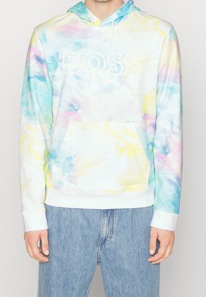 Osoba oblečená v světle modré, žluté a růžové batikované mikině s nápisem "BOSS" a modrých džínách, stojící s uvolněnýma rukama.