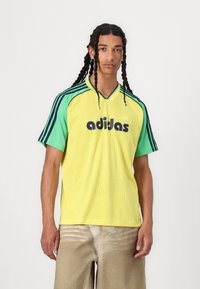 Mrežasta atletska majica v svetlo rumeni barvi z zelenimi akcenti. Ima ovratnik v obliki črke V, kratke rokave in izstopajoči črni logotip "adidas".