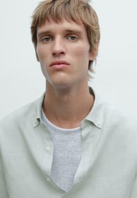 Chemise vert clair à boutons sur un t-shirt gris. La chemise présente un tissu doux avec un col décontracté et des boutons visibles. Fond neutre.