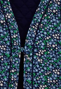 Námornícky prešívaný odev s kvetinovým vzorom v svetlomodrej a bielej farbe, s jemnými zelenými akcentami. Zapína sa na viazanie vpredu.