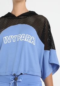 Blå kortärmad hoodie med svarta nätärmar, med "IVY PARK"-text i vitt. Dragsnöret i fållen justerar passformen.